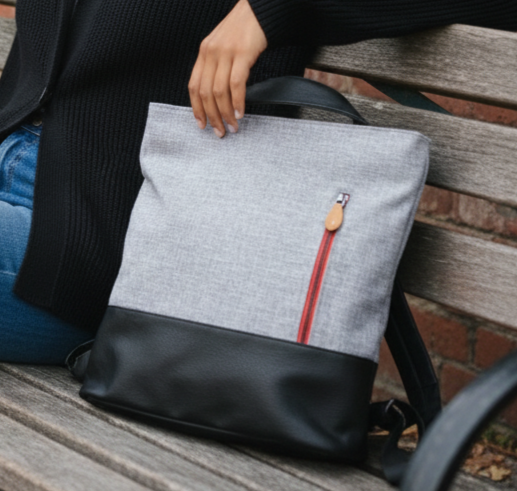 A gray and black tote back with a zipper pocket on the outside sits on a bench next to a person.
