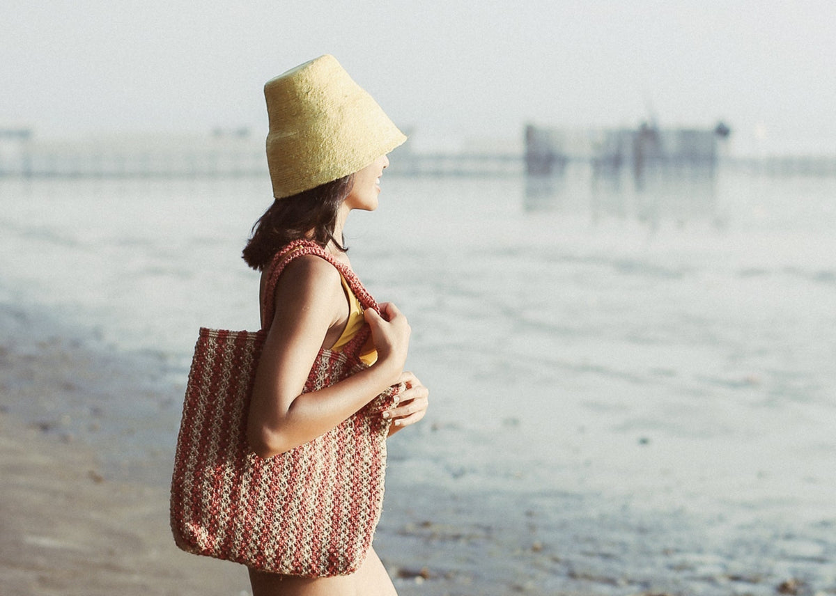 Woman walking on a beach wearing a yellow hat and carrying a red and beige patterned tote