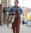 Woman walking on a sidewalk wearing a denim jacket and brown pants, holding a gold puffer laptop tote.