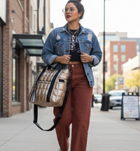 Woman walking on a sidewalk wearing a denim jacket and brown pants, holding a gold puffer laptop tote.