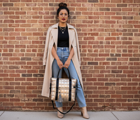 Woman in a beige coat and blue jeans holding a gold puffer laptop tote against a brick wall.