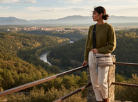 Woman standing on a lookout point overlooking a scenic landscape with a river and mountains holding a gold tote