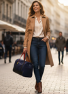 Woman in a tan coat and blue jeans walking on a city street with a navy bag.