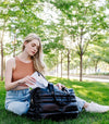 Woman sitting on grass reading a book with a black puffer weekender tote bag beside her