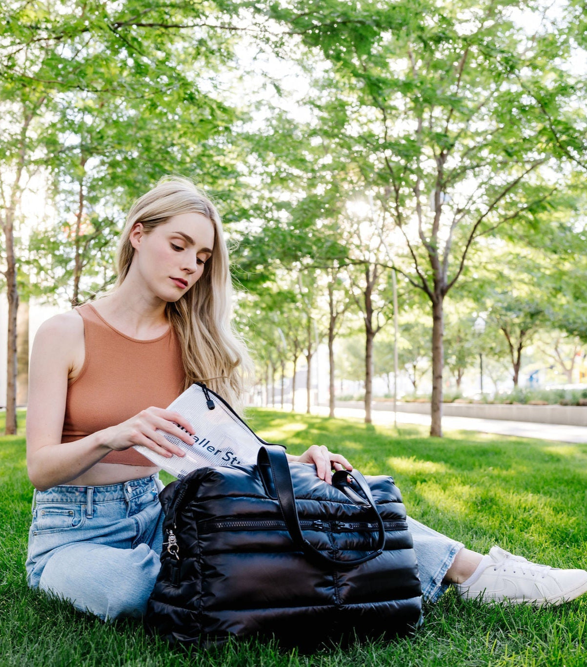 Woman sitting on grass reading a book with a black puffer weekender tote bag beside her