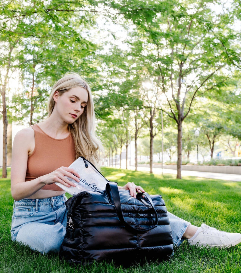 Woman sitting on grass reading a book with a black puffer weekender tote bag beside her