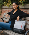 Woman sitting on a bench with a minimalist black and gray convertible backpack tote bag, surrounded by greenery and a brick wall.