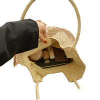 Hand opening a tan woven tote with cosmetic items on a white background