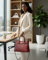 Woman in a beige suit holding a burgundy tote handbag in an office setting with large windows and a plant.