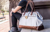 Person holding a stone gray weekender tote with brown handles and trim on a city street.