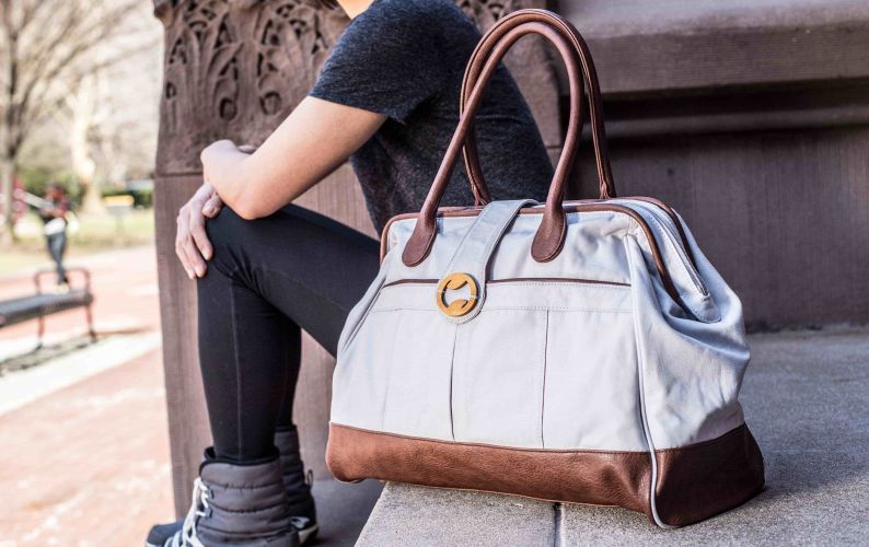 Person holding a stone gray weekender tote with brown handles and trim on a city street.