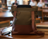 Green backpack convertible tote bag on a wooden table in a library setting with bookshelves and people in the background.