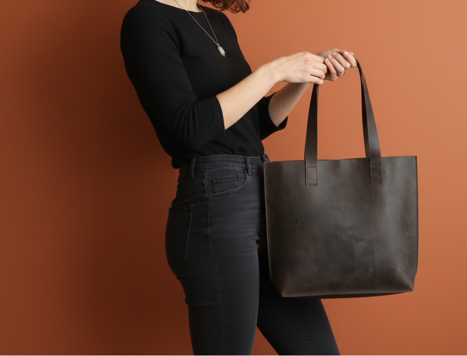 Woman holding a dark brown leather tote bag in an autumn setting with trees and fallen leaves.